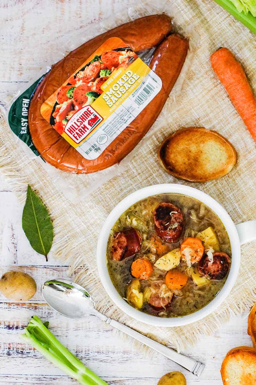 An overhead view of a package of smoked sausage next to a bowl of smoked sausage stew surrounded by vegetables on a white board. 