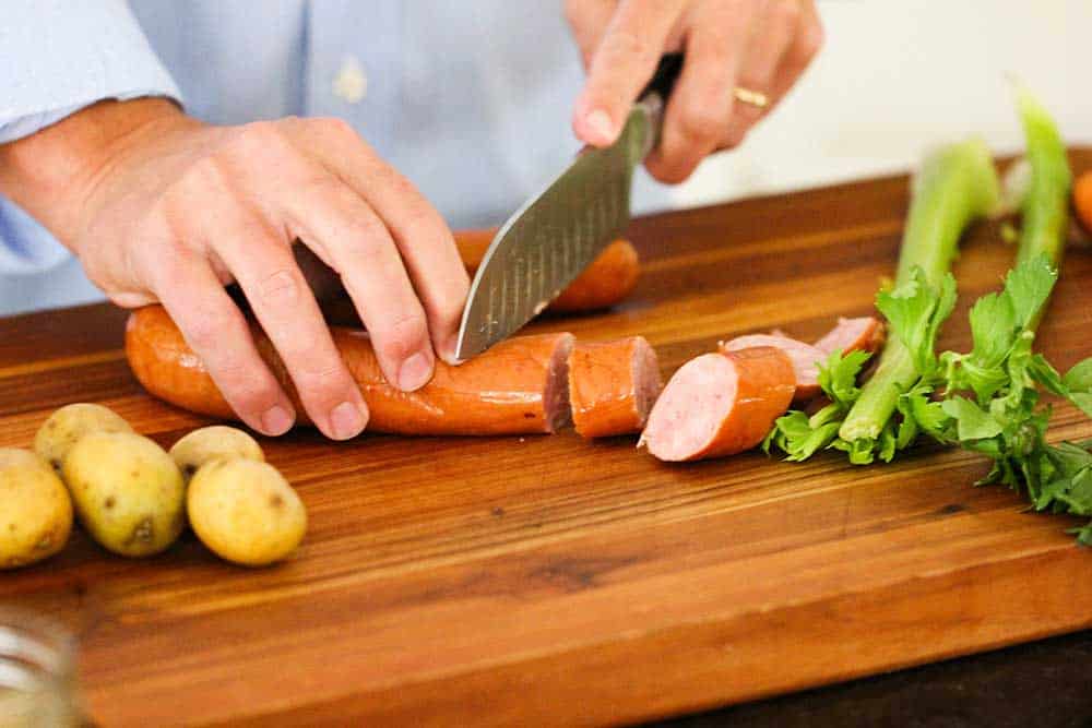 Two hands using a chef's knife to cut thick slices of smoked sausage and potatoes on a cutting board. 