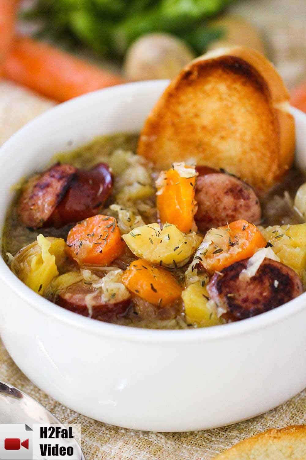 A large white bowl filled with smoked sausage stew and a piece of toasted bread stuck in the side with carrots in the background. 