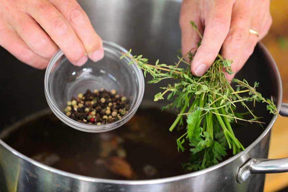 Two hands holding a small bowl of peppercorns and a bunch of herbs over a large stock pan for roasted chicken stock. 