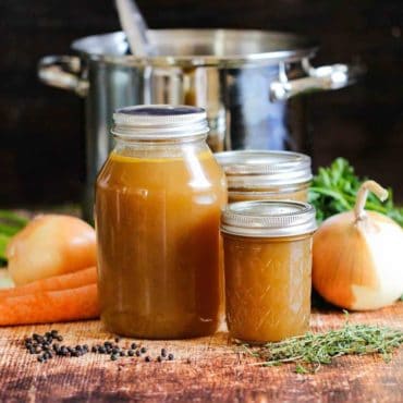 Several jars filled with roasted chicken stock in front of a large stock pan with vegetables and herbs nearby.