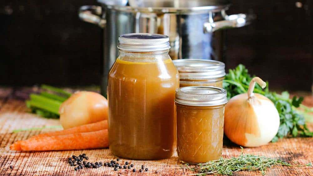 Three varied-sized jars holding roasted chicken stock in front of a large stock pan next to vegetables and herbs. 