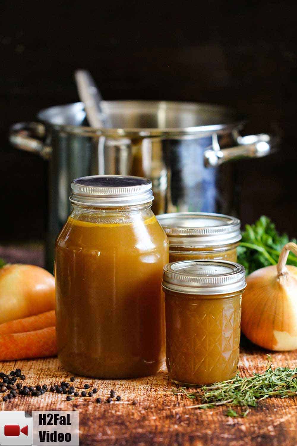 Three jars filled with roasted chicken stock with a stock pot and vegetables around them. 