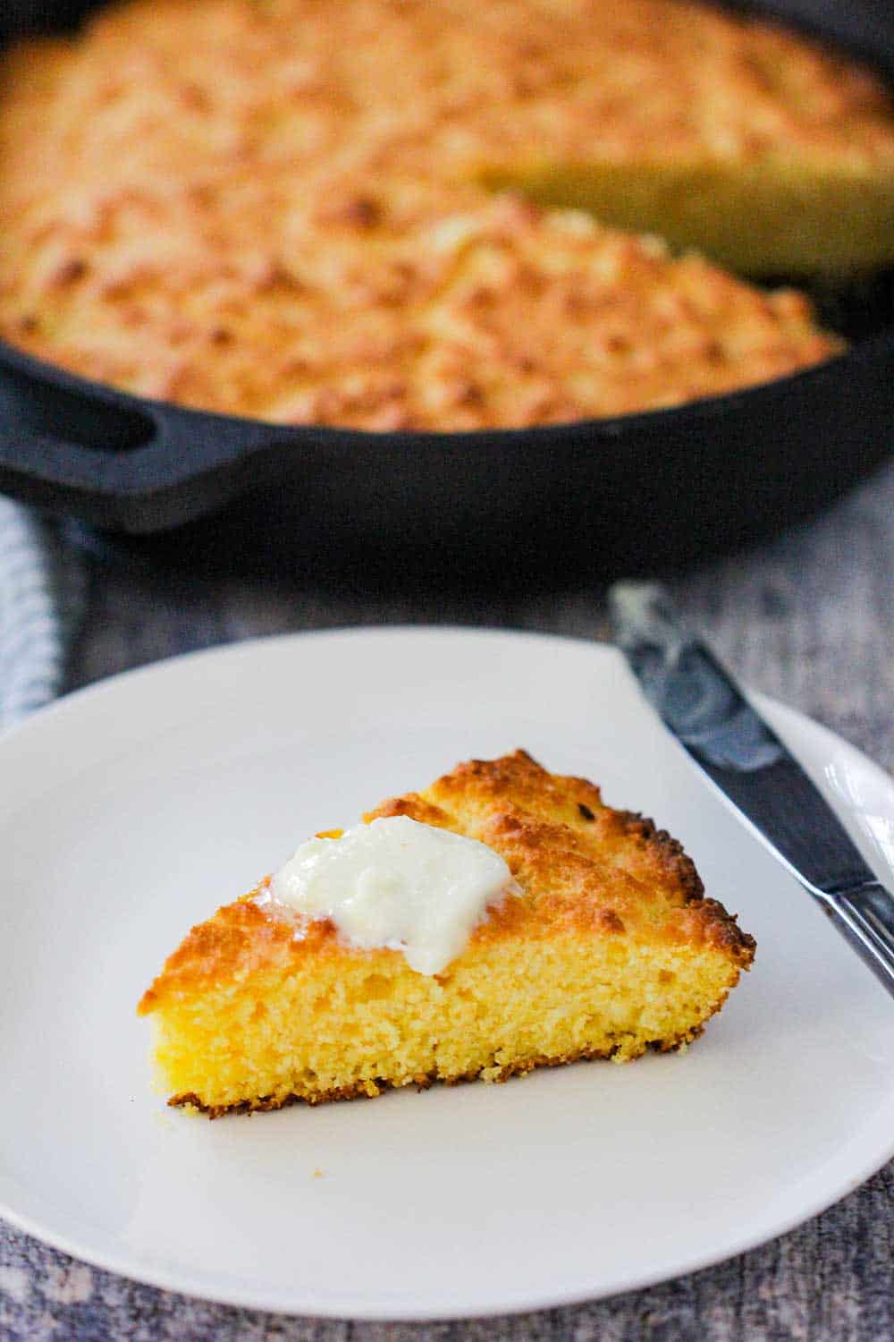 A slice of homemade cornbread with a pad of butter on top next to a skillet of cornbread. 