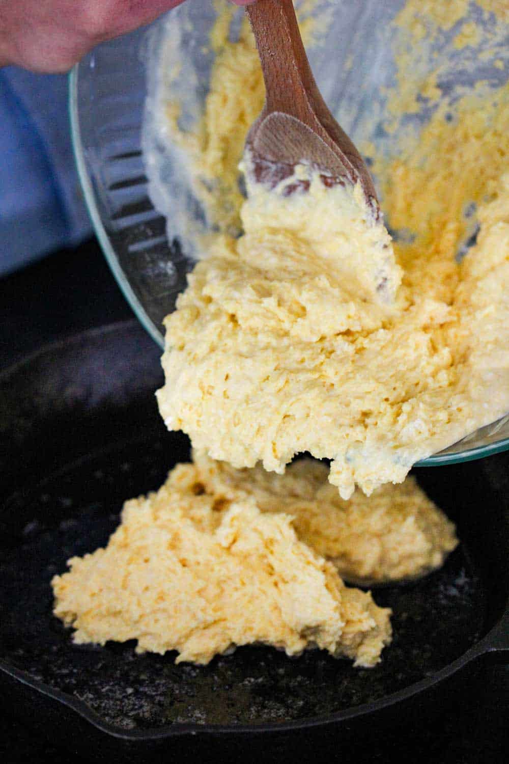 Cornbread batter being poured into a cast iron skillet from a glass bowl and a wooden spoon. 