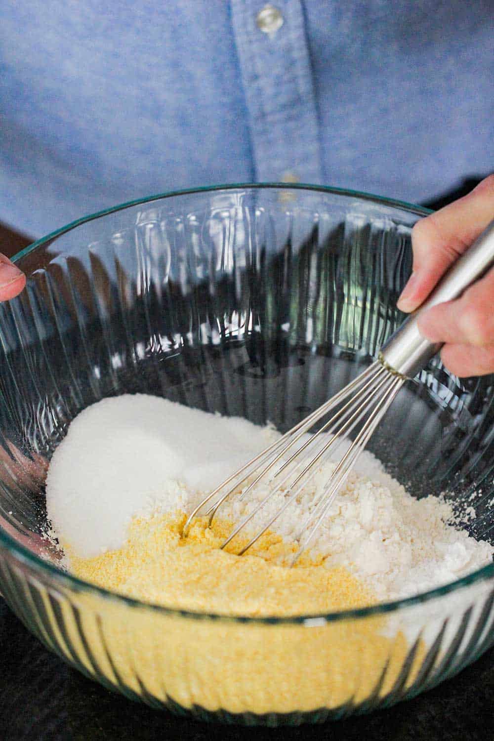 A hand using a whisk to combine cornmeal, flour, salt and baking powder in a large glass bowl. 