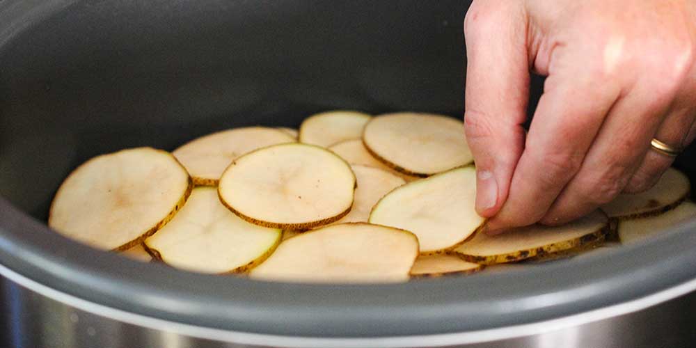 A hand adding a layer of sliced potatoes in a slow cooker. 