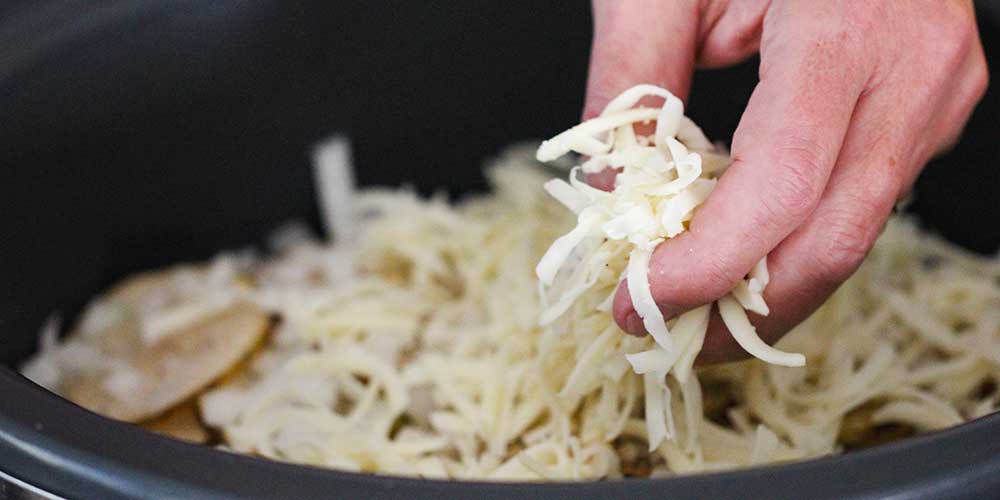 A hand dropping shredded Swiss cheese into a slow cooker lined with sliced potatoes. 