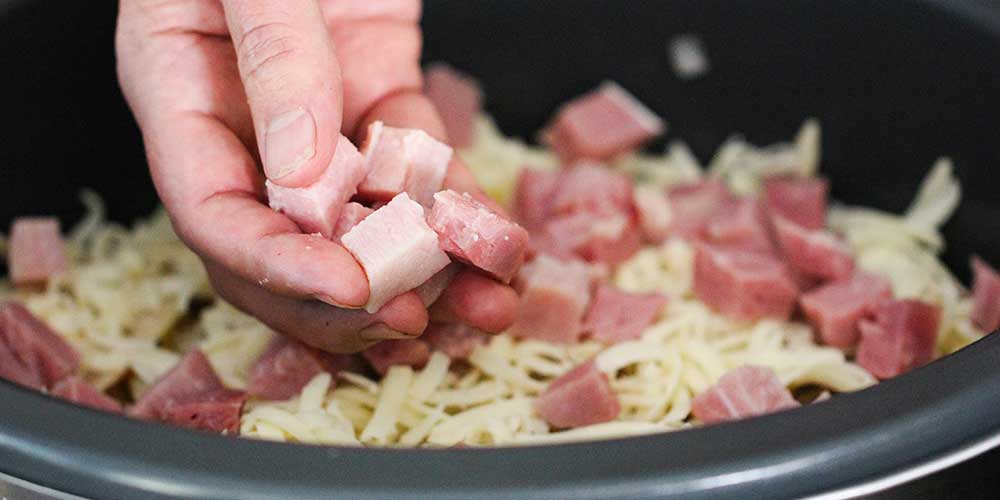 A hand dropping cubed ham into a Crock Pot for Slow-Cooker Ham and Potatoes au Gratin. 