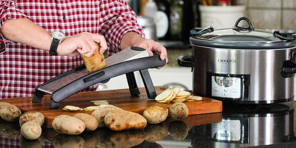 A hand slicing a russet potato on a mandolin next to a large Crock-Pot. 