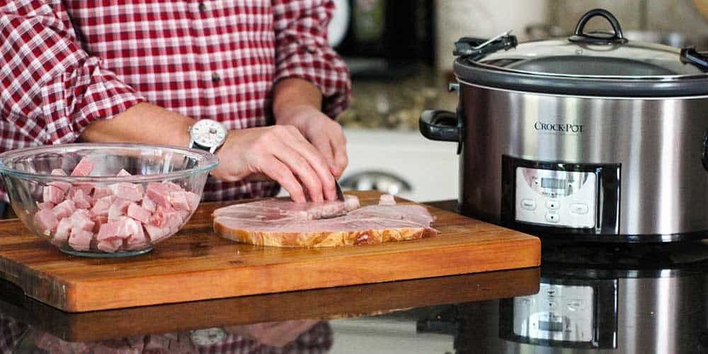 Two hands cutting a ham steak on a cutting board next to a large slow-cooker. 