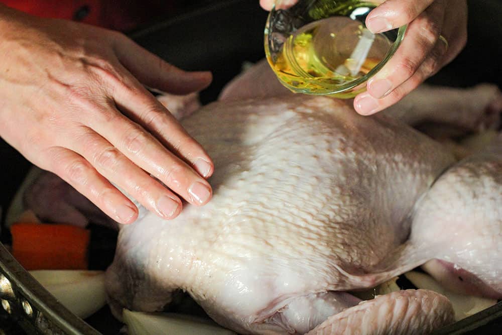 Two hands pouring oil onto an uncooked turkey in a roasting pan. 