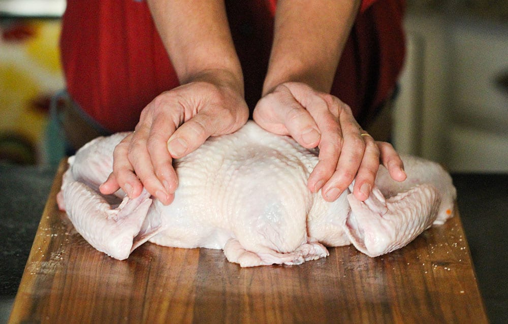 Two hands pressing down on an uncooked turkey that has been spatchcocked. 