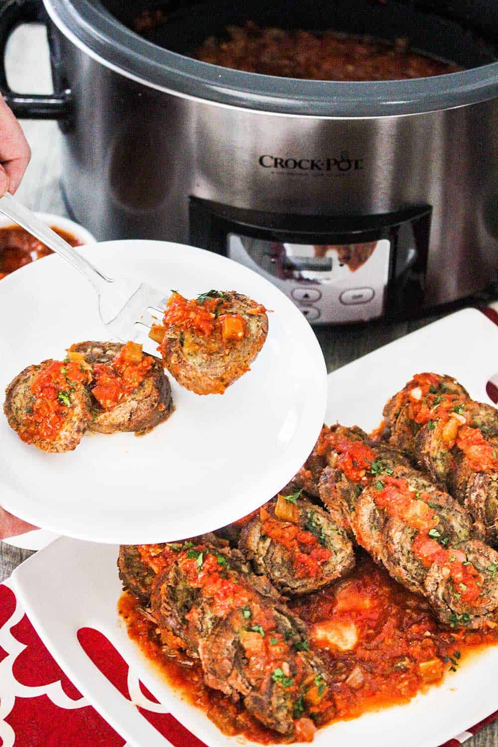 A hand holding a serving of beef braciole on a white plate next to a serving dish and a slow cooker. 