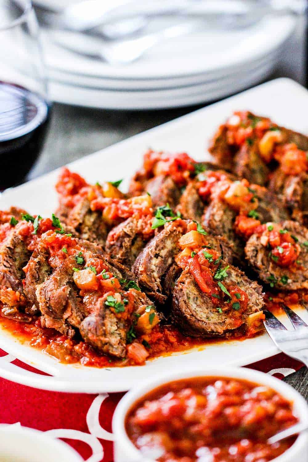 A white platter holding sliced slow cooker beef braciole with a bowl of marinara next to it. 