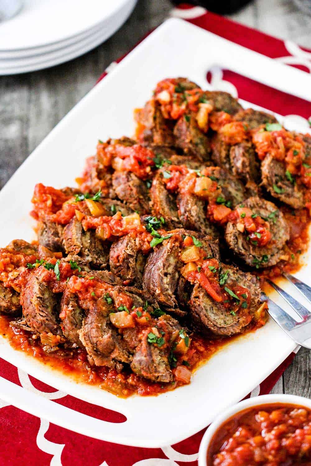 A close up view of slow cooker beef braciole on a white platter next to a bowl of marinara sauce.