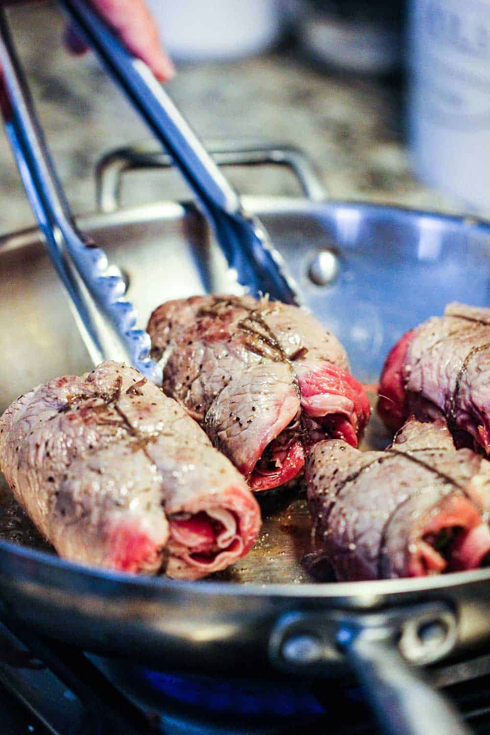 A pair of tongs holding slow cooker beef braciole in a large skillet. 