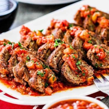 A close up view of slow cooker beef braciole on a white platter next to a bowl of marinara sauce.