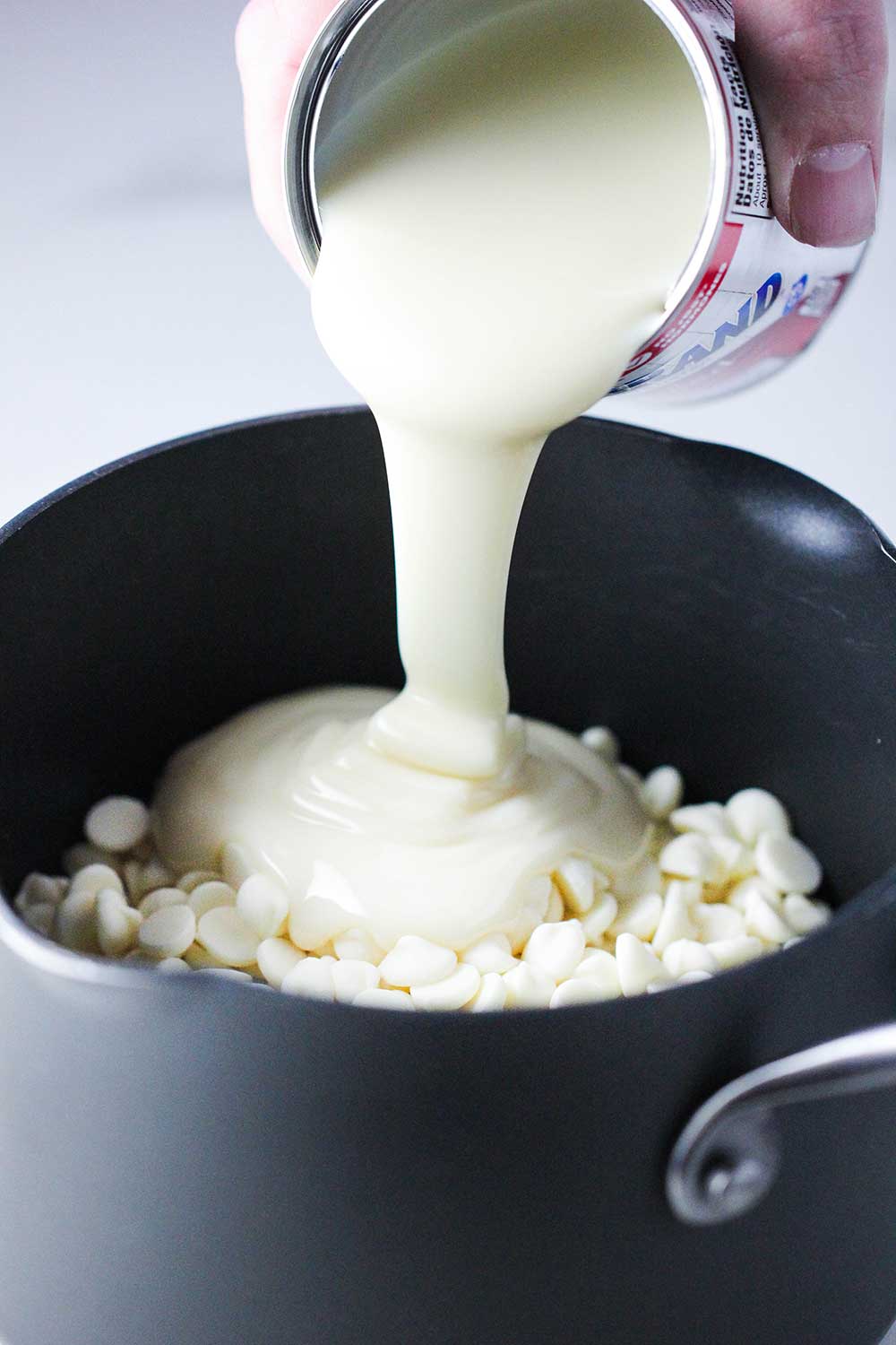 A hand pouring a can of condensed milk into a pan of white chocolate. 