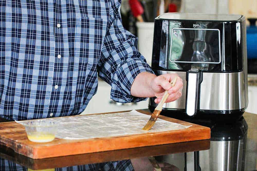 A hand using a brush to apply melted butter onto phyllo dough next to an air fryer. 