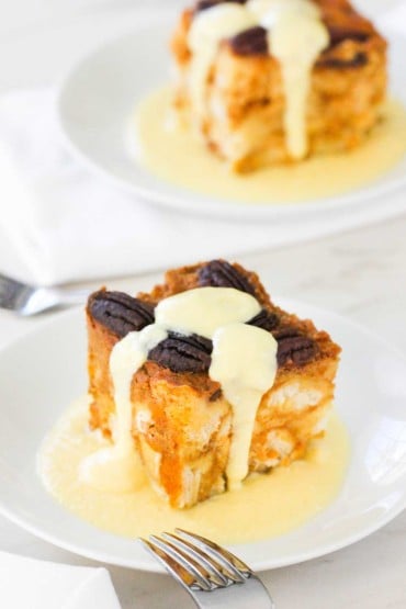 A piece of pumpkin bread pudding with vanilla custard poured over the top on a white plate.