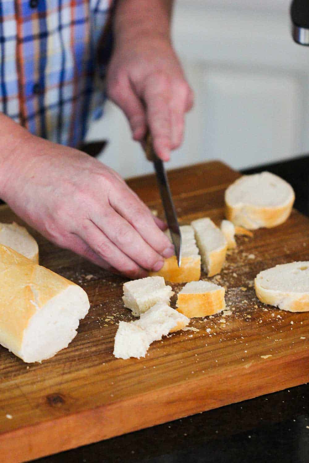 Two hands using a knife to cut a French loaf of bread into cubes. 