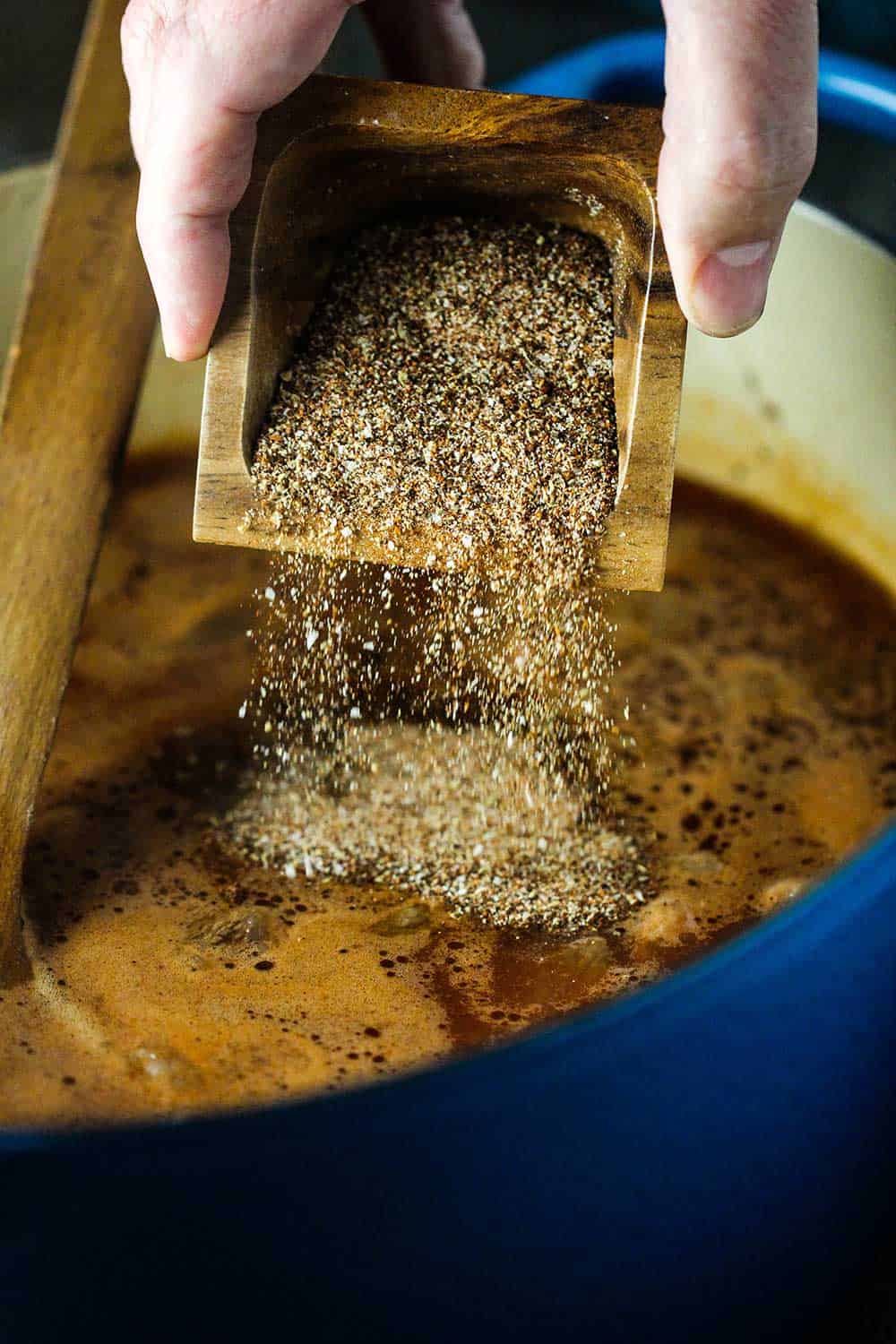 A hand dumping a square container of chili seasoning into a pot of meat and sauce.