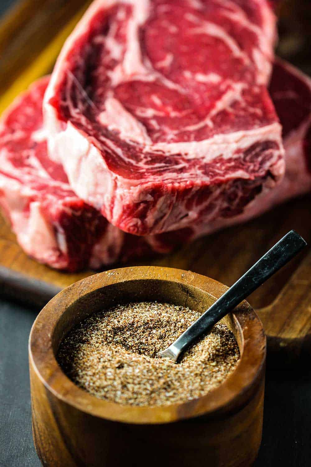 Two large boneless ribeye steaks on a cutting board next to a wooden bowl of spices.