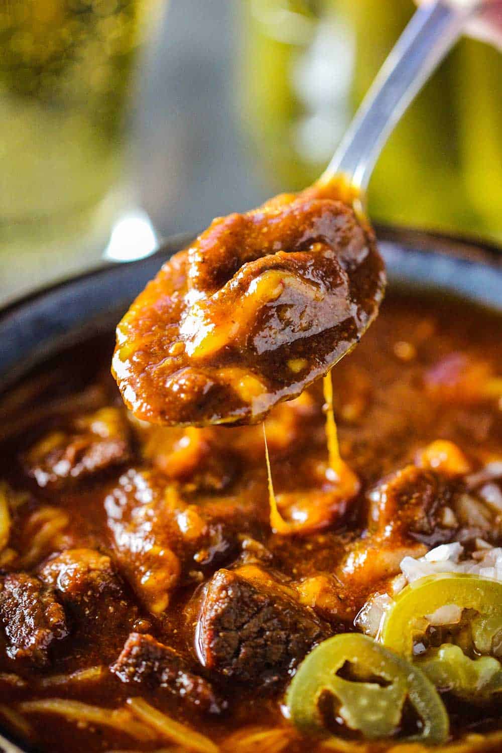 A spoon lifting a helping of Texas-style chili from a bowl of the chili with cheese dripping off the sides.