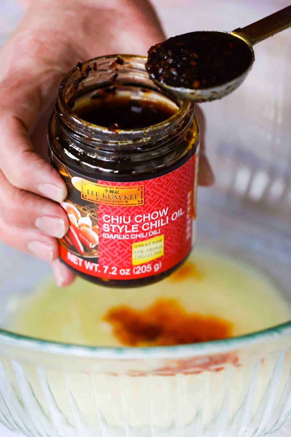 One hand holding a jar of chili oil with another hand holding a tablespoon of the sauce over a bowl of melted butter. 