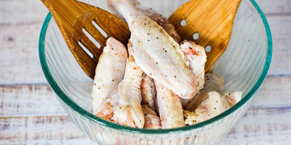 A small glass bowl of uncooked chicken wings with two wooden spoons in the bowl. 