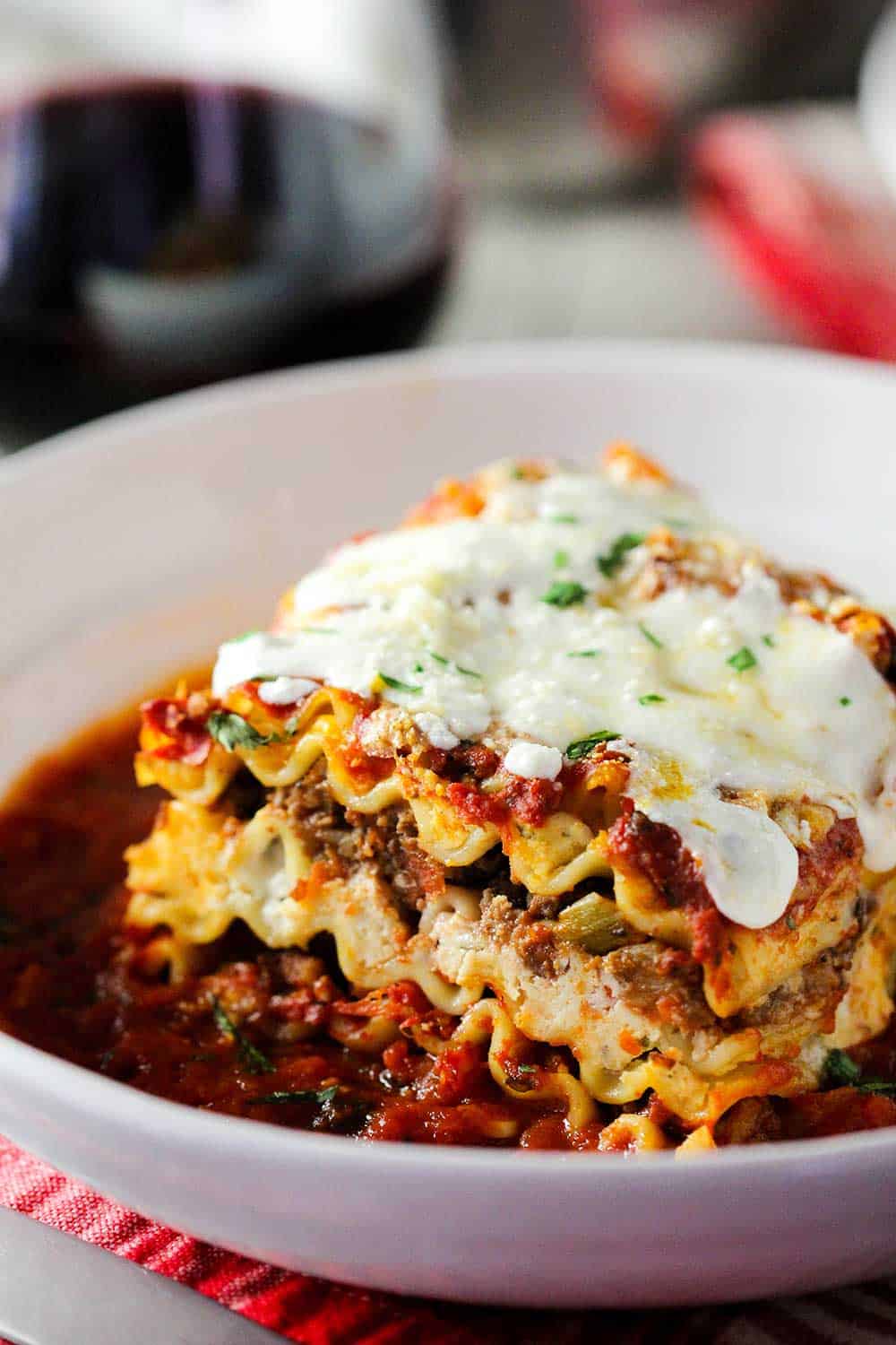 A white bowl of classic meat lasagna next to a glass of red wine. 