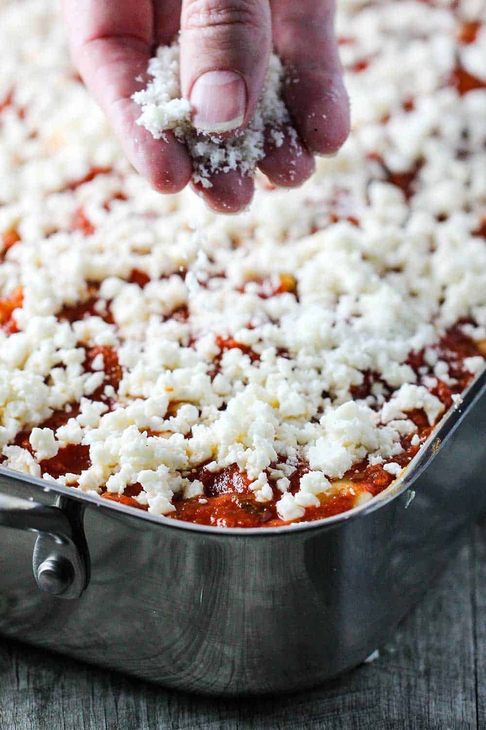 A hand sprinkling mozzarella cheese on top of a full lasagna pan of lasagna. 