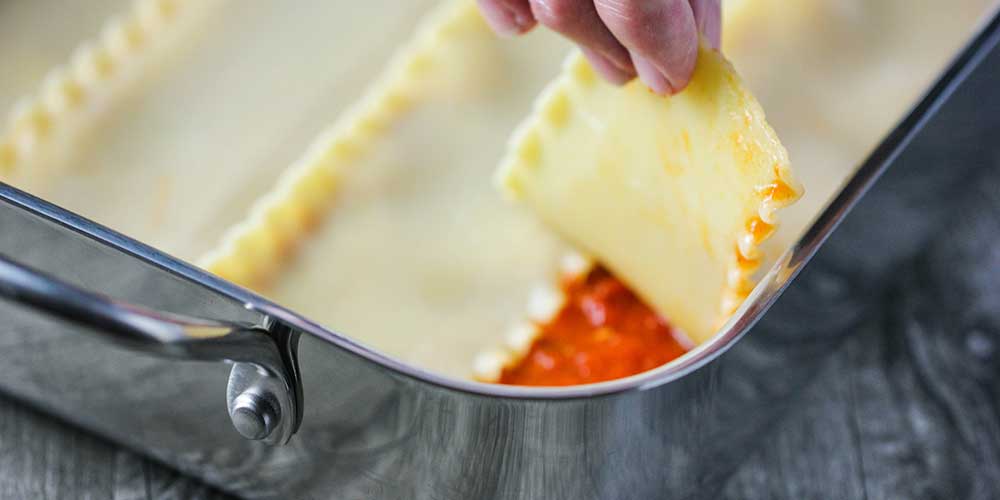 A hand laying lasagna noodles over a layer of marinara in a lasagna dish. 