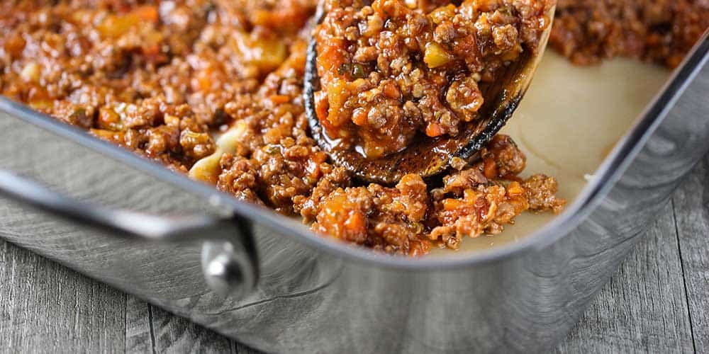 A wooden spoon layering meat sauce of noodles in a lasagna dish. 