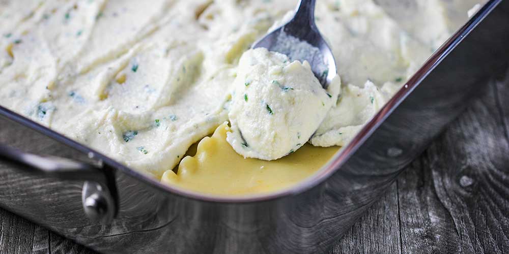 A spoon spreading ricotta cheese over noodles in a lasagna pan. 