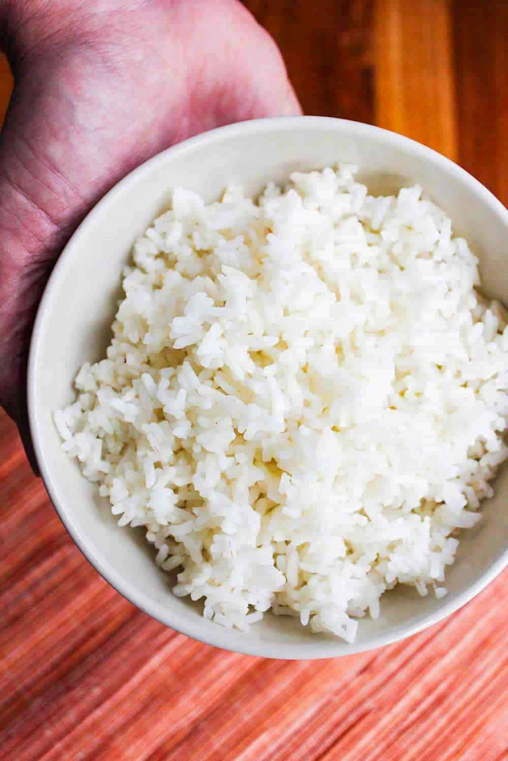 An overhead view of a hand holding a white bowl of cooked rice. 