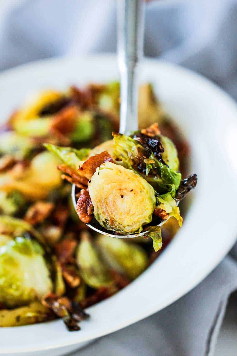 A large silver spoon holding a serving of maple-roasted Brussels sprouts with bacon and pecans over a dish of the same. 