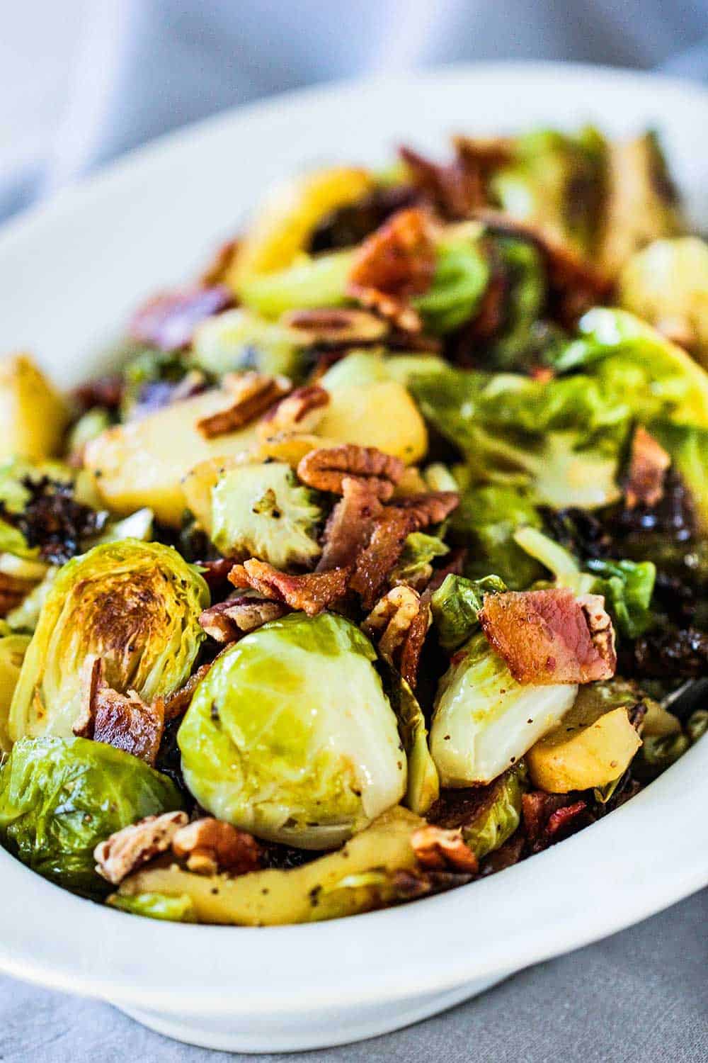 A white serving bowl containing maple-roasted Brussels sprouts with bacon and pecans next to a grey cloth and spoon. 