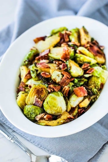 A white serving bowl containing maple-roasted Brussels sprouts with bacon and pecans next to a grey cloth and spoon.
