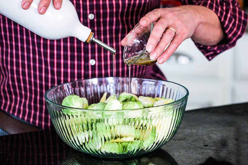 A person pouring bacon grease from a small dish in one hand, and pouring olive oil from a container with the other hand into a bowl of uncooked Brussels sprouts. 