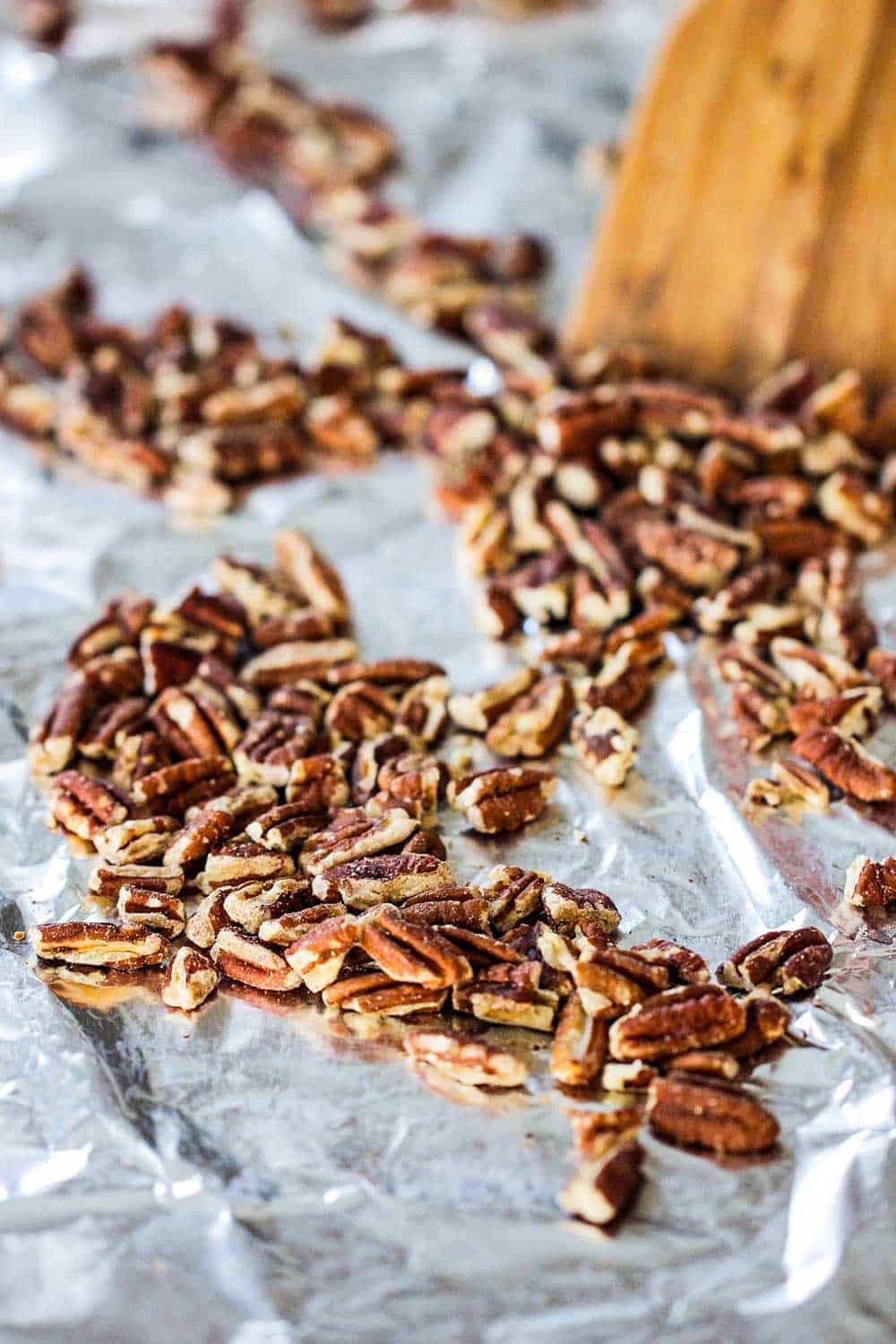 A baking sheet lined with foil with pecans on it and a wooden spoon scooping them. 