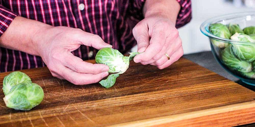 Two hands pulling away loose leaves from a Brussels sprout on a wooden cutting board. 