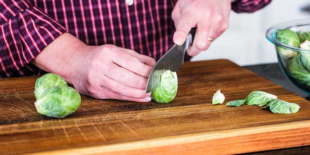 Two hands holding a large knife cutting a Brussels sprout down the middle on a cutting board. 