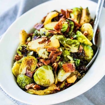 A white serving bowl containing maple-roasted Brussels sprouts with bacon and pecans next to a grey cloth and spoon.