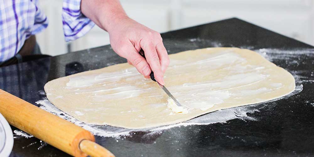 A hand spreading softened butter on a rectangle of pastry dough.