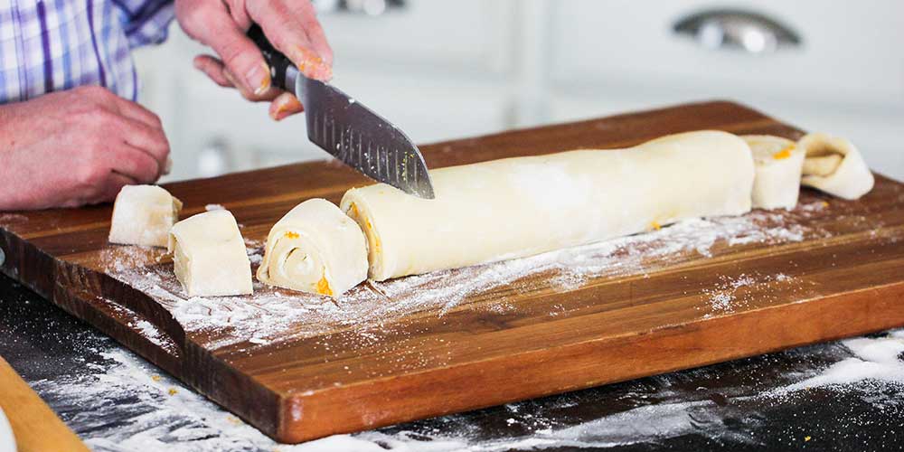 A hand using a large chef's knife to slice sweet rolls from rolled dough.