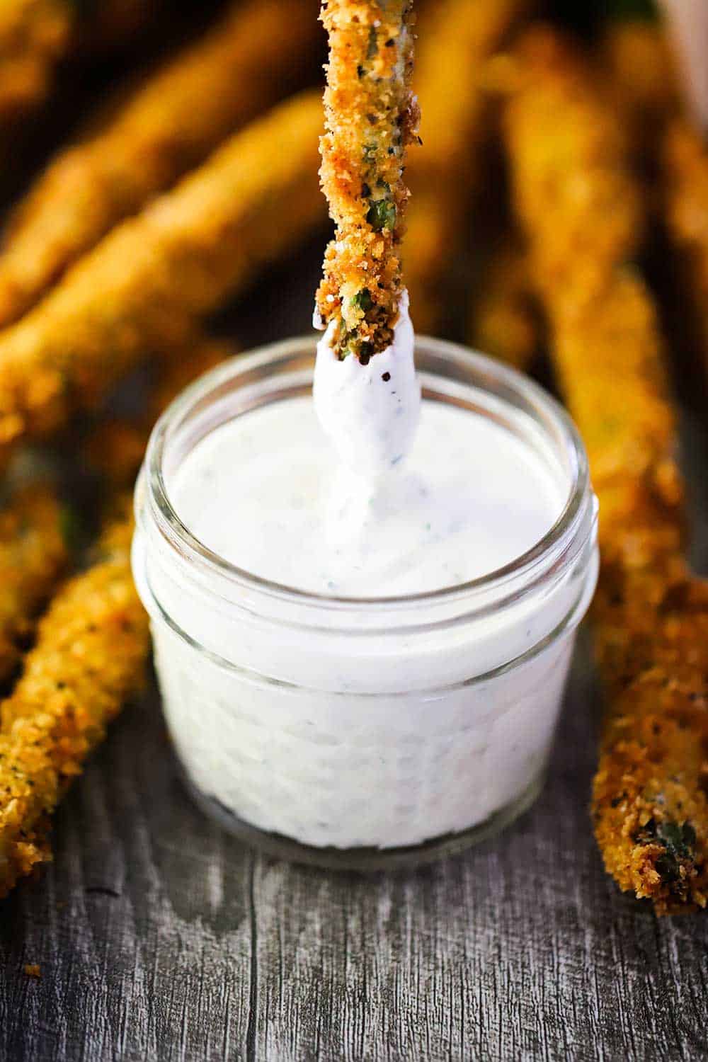 A small jar of ranch dressing in the middle of a bunch of fried asparagus with one being lowered into the jar. 
