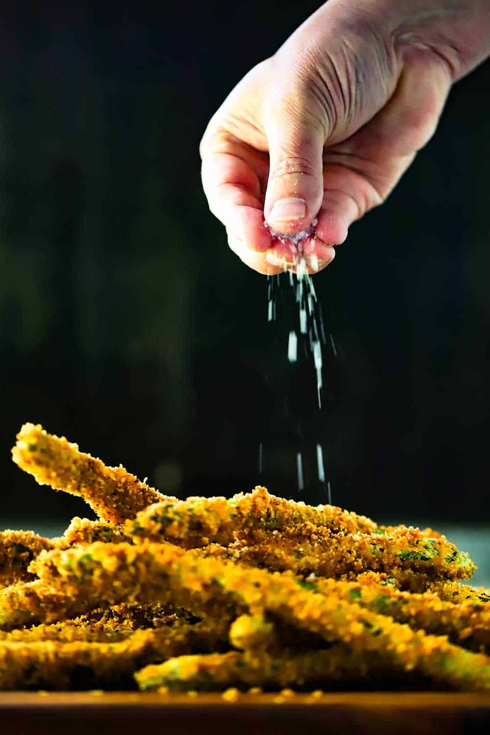 A hand sprinkling coarse sea salt of a pile of fried asparagus. 