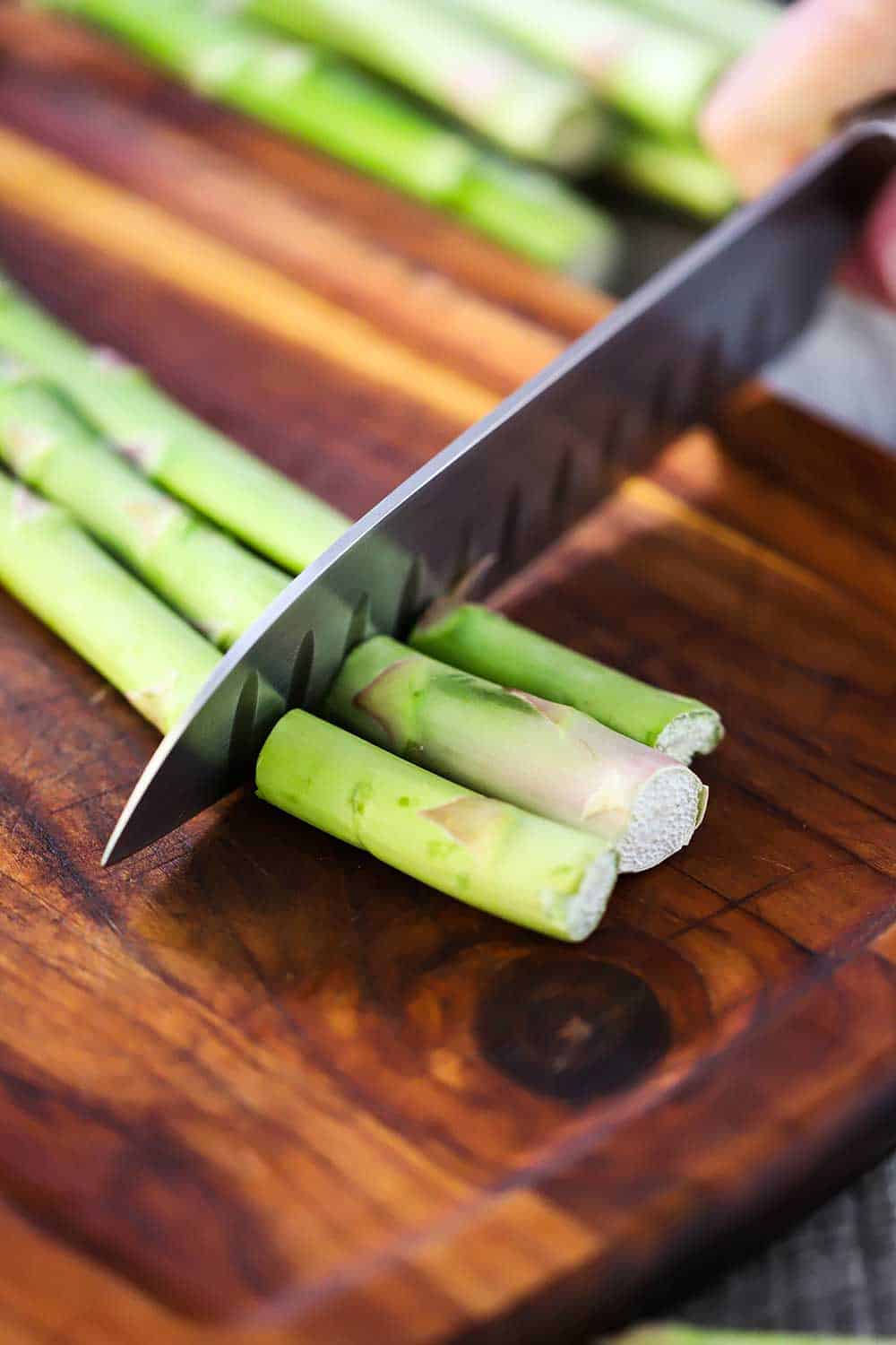 A hand using a chef's knife to cut the ends of of three asparagus stalks on a cutting board. 